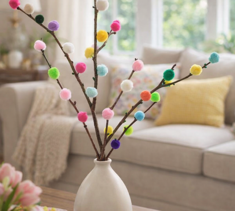 Farbige Pompons zieren mehrere Zweige in einer schlichten Vase auf einem Tisch, im Hintergrund ein gemütliches Sofa mit Kissen. | © Generiert mit KI ( Erstellt mit ChatGPT)