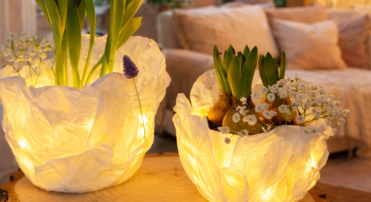 Zwei leuchtende Tulpen aus Butterbrotpapier stehen mit Frühlingsblumen auf einer Baumscheibe in gemütlich beleuchtetem Wohnzimmer.