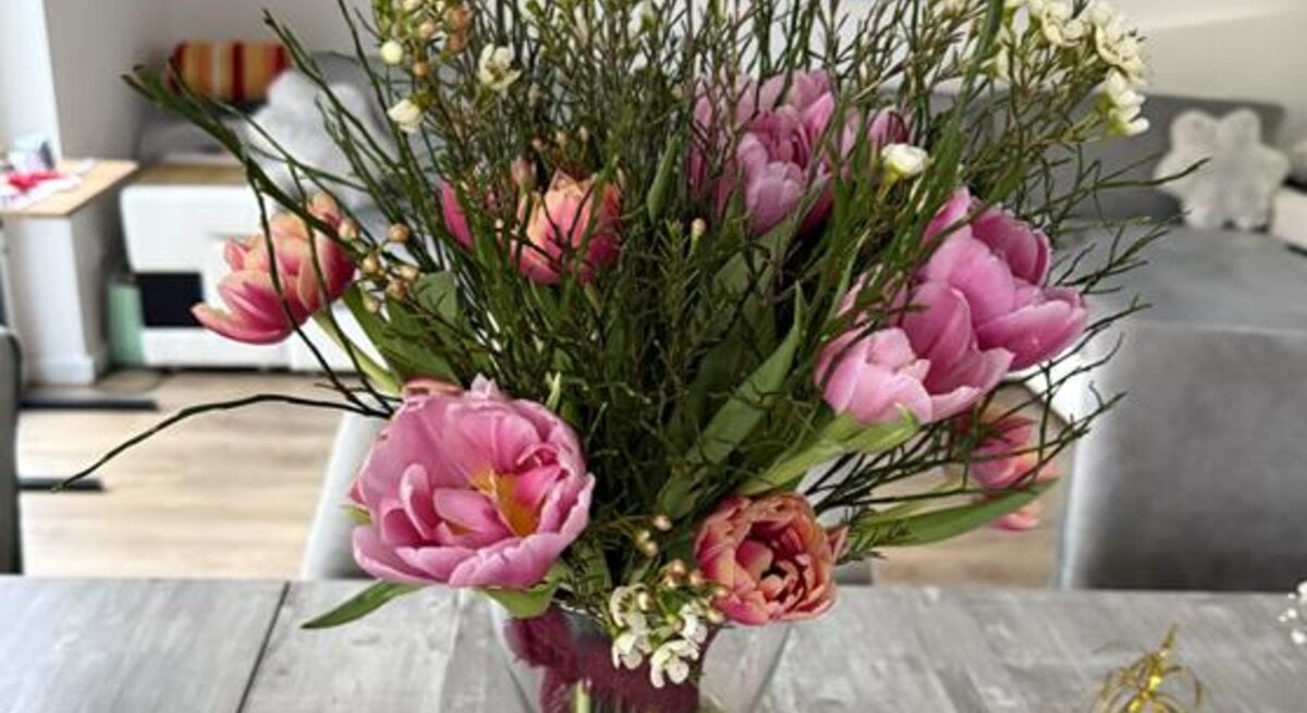 Ein bunter Frühlingsstrauß mit rosa Tulpen und kleinen weißen Blüten steht in einer Vase auf grauem Holztisch.