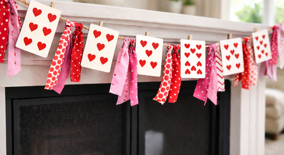 Spielkarten mit roten Herzen hängen an einer Girlande aus rosa-roten Stoffstreifen über einem hellen Kamin.