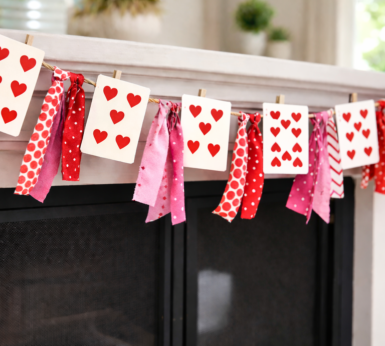 Spielkarten mit roten Herzen hängen an einer Girlande aus rosa-roten Stoffstreifen über einem hellen Kamin. | © Generiert mit KI (Erstellt mit ChatGPT) 