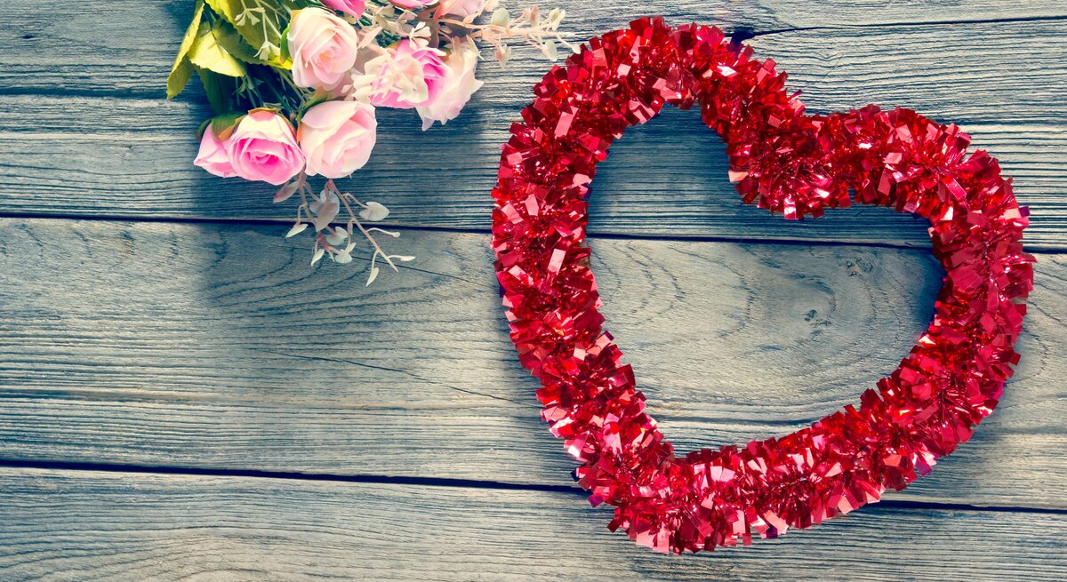 Ein Herz- Kranz aus roter Glitzergirlande liegt neben zarten rosa Rosen auf verwittertem Holztisch.