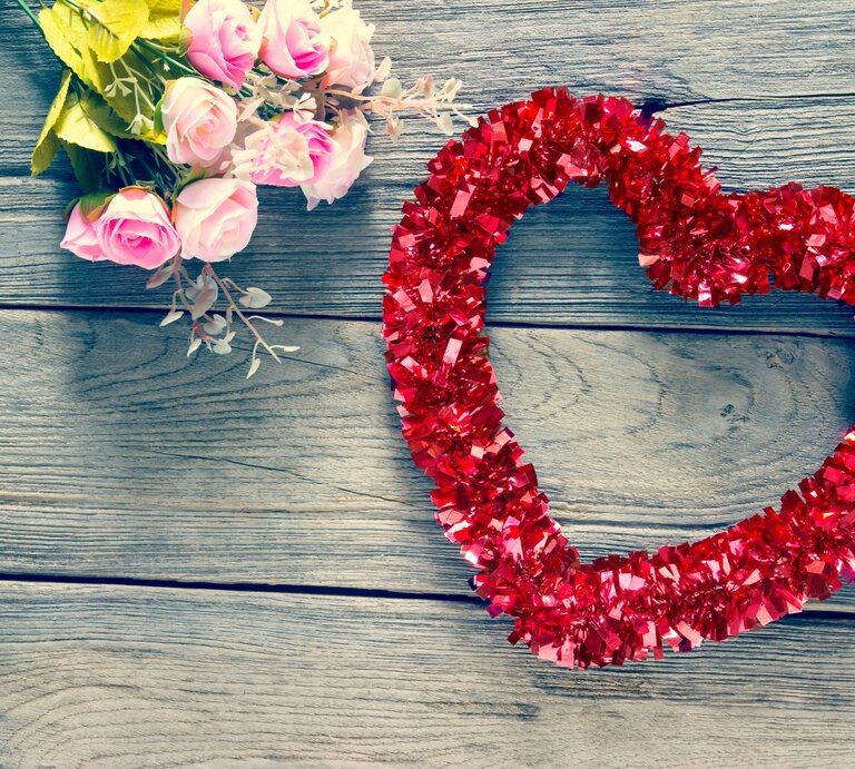 Ein Herz- Kranz aus roter Glitzergirlande liegt neben zarten rosa Rosen auf verwittertem Holztisch. | © Shutterstock/warat42