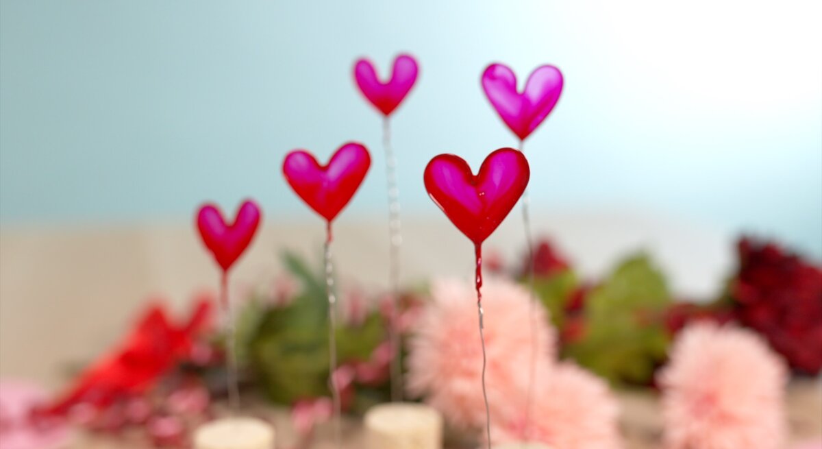 Herzförmige Deko auf Draht vor pastellblauem Hintergrund und verschwommenem Blumenarrangement aus Rosa und Rot.