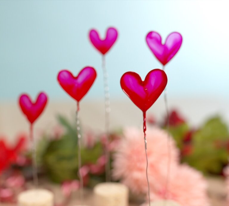 Herzförmige Deko auf Draht vor pastellblauem Hintergrund und verschwommenem Blumenarrangement aus Rosa und Rot. | © Bild der Frau 