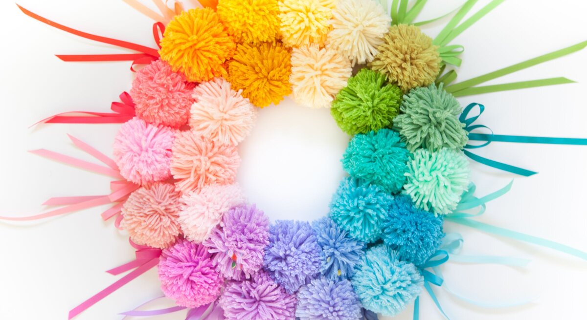 Regenbogenfarbener Pompon-Kranz aus weicher Wolle mit farbigen Geschenkbändern auf strahlend weißem Hintergrund arrangiert.