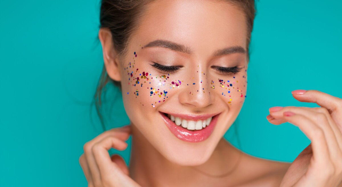 Konfetti Kostüme Damen basteln: Junge Frau mit glänzenden Lippen lächelt verträumt, bunte Glitzerpunkte zieren ihre Wangen vor türkisfarbenem Hintergrund.