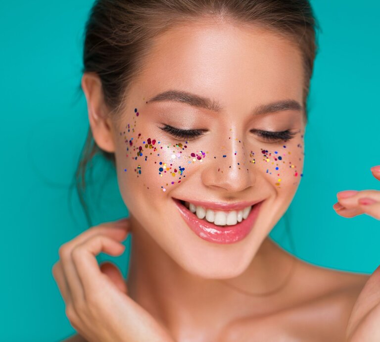 Konfetti Kostüme Damen basteln: Junge Frau mit glänzenden Lippen lächelt verträumt, bunte Glitzerpunkte zieren ihre Wangen vor türkisfarbenem Hintergrund. | © Shutterstock/Vikacita