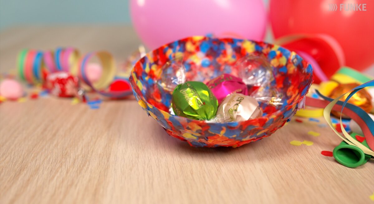 Farbenfrohe Konfetti-Schale mit Süßigkeiten auf hellem Tisch, daneben Schere, Papierstreifen und Luftballons in fröhlicher Partyatmosphäre.