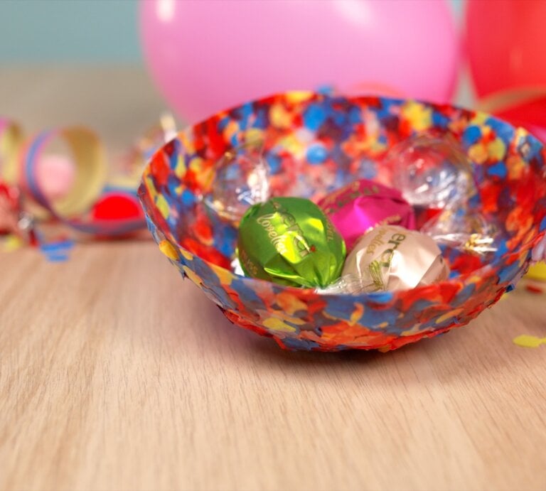 Farbenfrohe Konfetti-Schale mit Süßigkeiten auf hellem Tisch, daneben Schere, Papierstreifen und Luftballons in fröhlicher Partyatmosphäre. | © bildderfrau.de