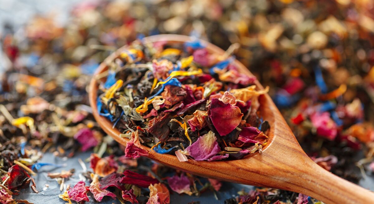 Bunte Blütenblätter und Teeblätter in kräftigen Farben auf einem Holzlöffel, lose verteilt auf dunklem Hintergrund.