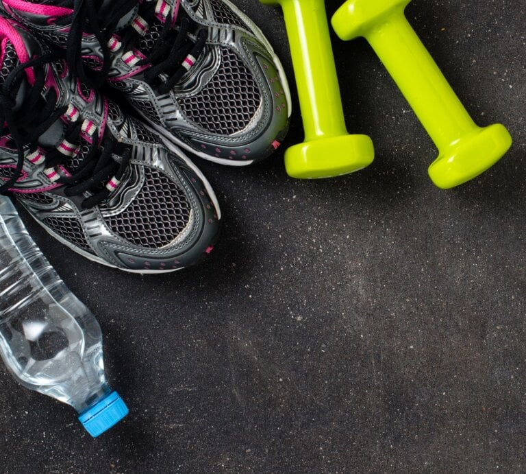 Zwei neongelbe Hanteln liegen neben grauen Sportschuhen mit pinken Details und einer Wasserflasche auf Asphaltboden. | © Shutterstock/Leszek Czerwonka