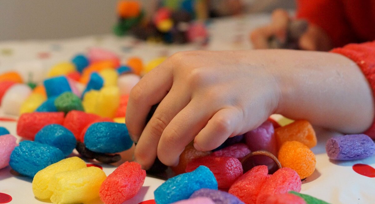 Kinderhand greift nach bunten Bastelchips aus Mais auf einem weißen Tisch mit roten Punkten im weichen Tageslicht.