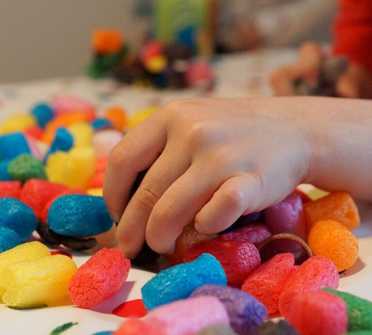 Kinderhand greift nach bunten Bastelchips aus Mais auf einem weißen Tisch mit roten Punkten im weichen Tageslicht. | © GettyImages/Cathérine