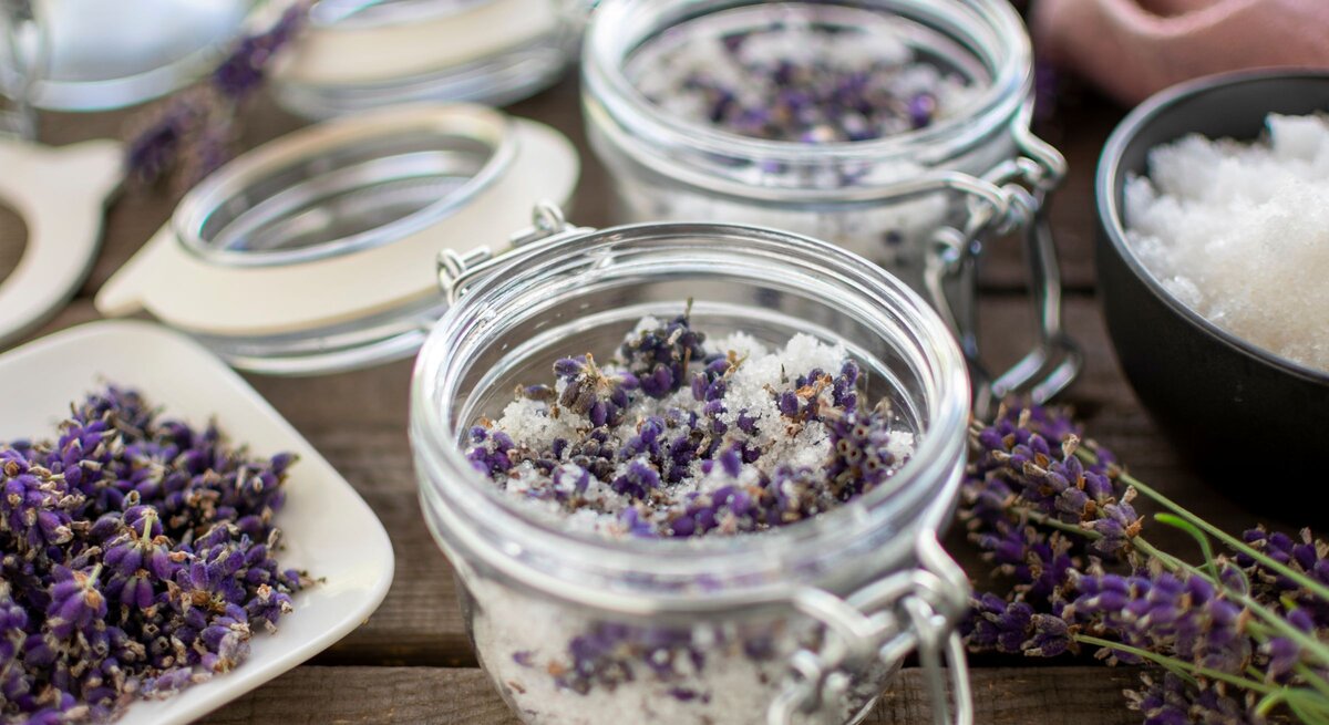 Ein Glas mit grobem Salz und getrocknetem Lavendel steht offen auf rustikalem Holz, daneben Blüten, Zucker und weitere Gläser.