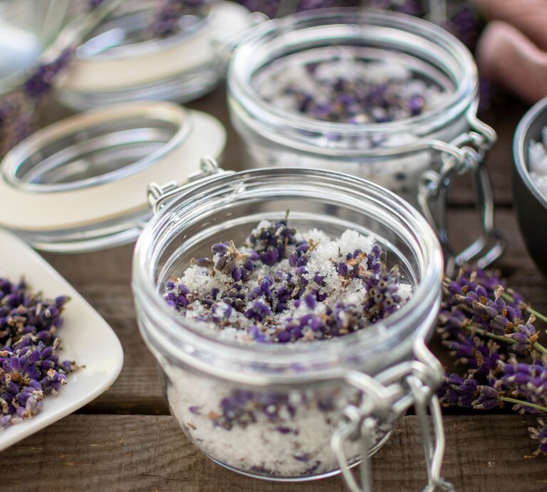 Ein Glas mit grobem Salz und getrocknetem Lavendel steht offen auf rustikalem Holz, daneben Blüten, Zucker und weitere Gläser. | © AdobeStock/Stefanie