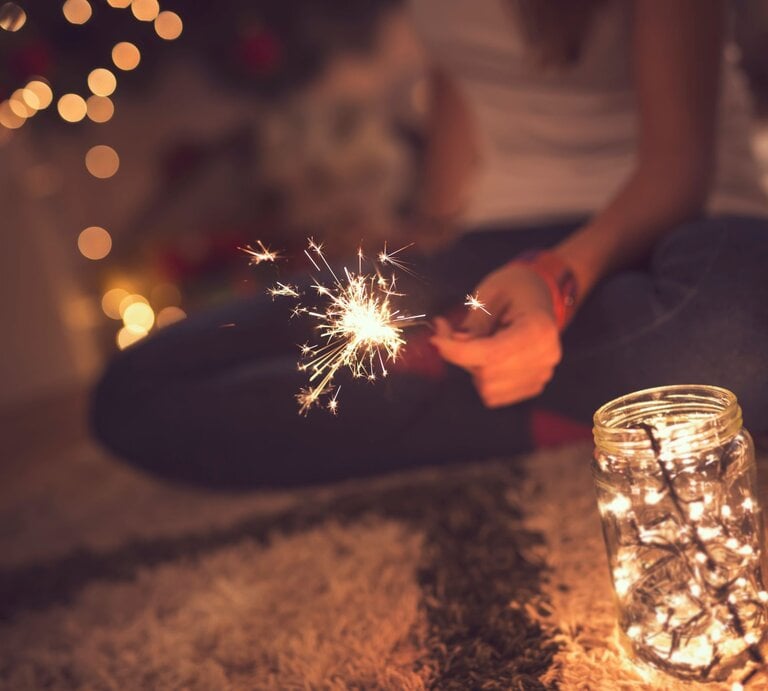 Junge Frau mit Wunderkerze sitzt im Schneidersitz auf Teppich, neben ihr ein Windlicht mit warm leuchtender Lichterkette. | © GettyImages/vladans