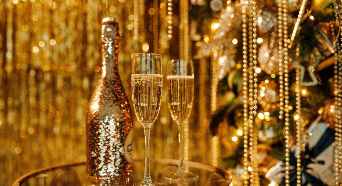 Zwei gefüllte Sektgläser und eine glitzernde Sekt-Flasche stehen vor goldenem Lametta und funkelndem Christbaumschmuck.