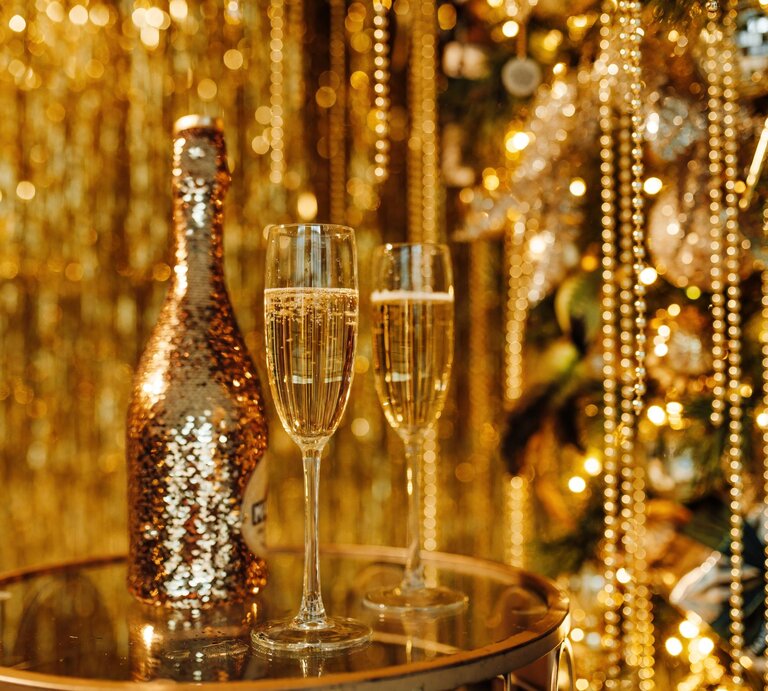 Zwei gefüllte Sektgläser und eine glitzernde Sekt-Flasche stehen vor goldenem Lametta und funkelndem Christbaumschmuck. | © Shutterstock/Olha Yefimova