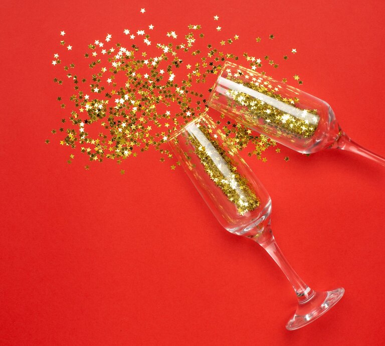 Zwei Sektgläser stoßen an, goldene Stern-Konfetti wirbeln sprühend über rotem Hintergrund durch die Luft. | © Shutterstock/KatrinaEra