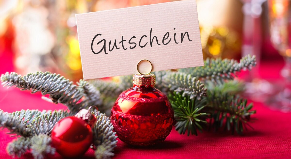 Rote Christbaumkugel mit Kärtchenaufsatz „Gutschein“ liegt auf Tannenzweig vor festlich verschwommenem Hintergrund.