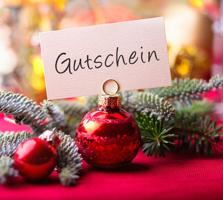 Rote Christbaumkugel mit Kärtchenaufsatz „Gutschein“ liegt auf Tannenzweig vor festlich verschwommenem Hintergrund. | © Shutterstock/Floydine