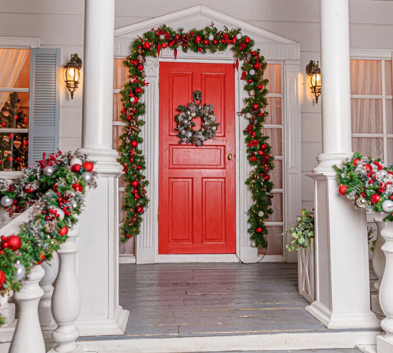 Weihnachtlich geschmückte Veranda mit roter Tür, grünen Girlanden und silbernem Kranz, im Fenster leuchtet ein Christbaum. | © Shutterstock/Julia Zavalishina