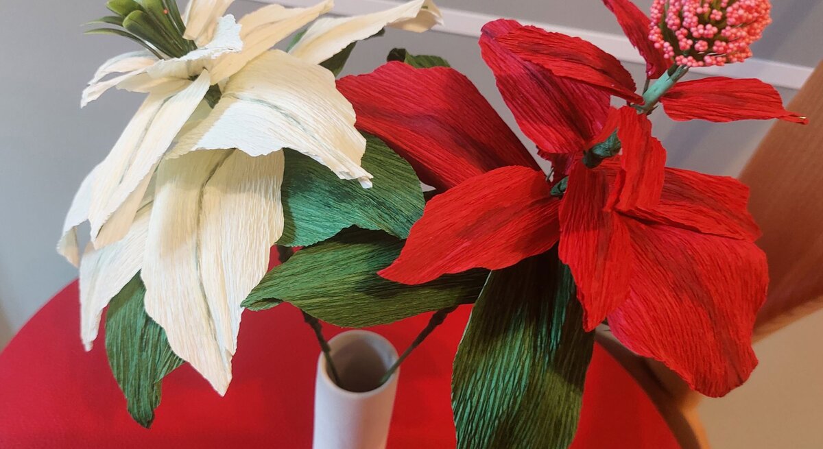 Zwei handgefertigte Krepppapierblumen, eine in Rot und eine in Weiß, mit grünen Blättern in eleganter Vase auf rotem Tisch vor grauem Hintergrund.