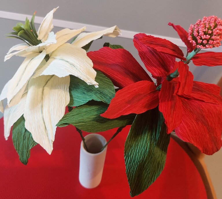 Zwei handgefertigte Krepppapierblumen, eine in Rot und eine in Weiß, mit grünen Blättern in eleganter Vase auf rotem Tisch vor grauem Hintergrund. | © Linda Pötzsch