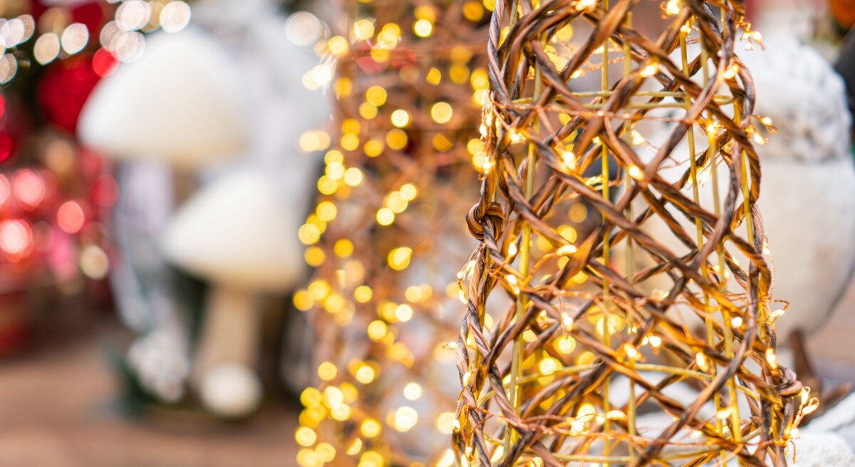 Geflochtener Drahtkegel mit warmweißen Lichterketten vor verschwommenem Weihnachtsdekor in Rot und Weiß.