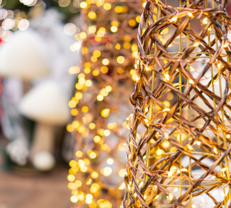 Geflochtener Drahtkegel mit warmweißen Lichterketten vor verschwommenem Weihnachtsdekor in Rot und Weiß. | © Shutterstock/ mr_tigga