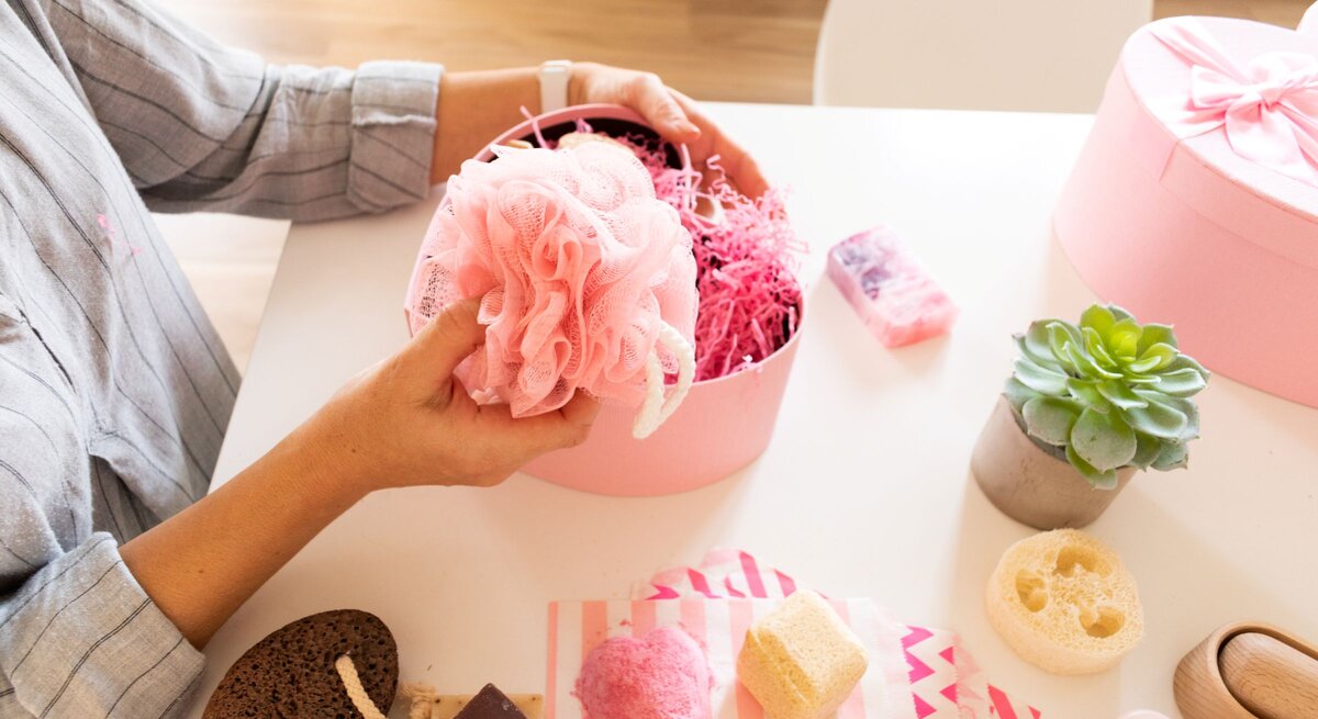 Frau in gestreifter Bluse hält rosa Duschschwamm über geöffneter Geschenkbox mit Pflegeprodukten auf hellem Tisch.