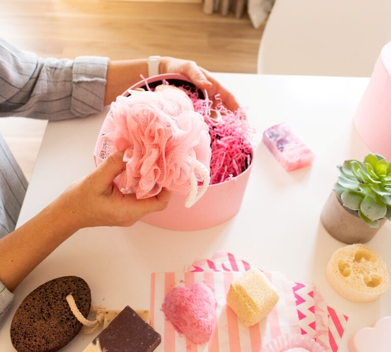 Frau in gestreifter Bluse hält rosa Duschschwamm über geöffneter Geschenkbox mit Pflegeprodukten auf hellem Tisch. | © Shutterstock/Kolpakova Daria