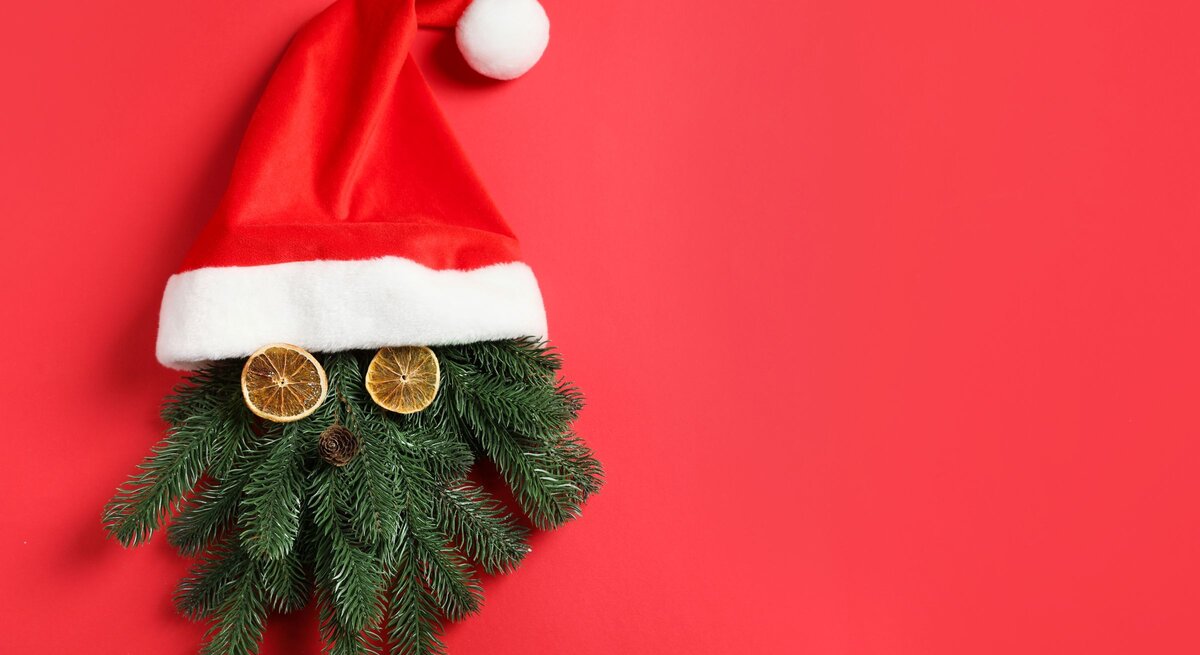 Tannenzweige mit getrockneten Orangenscheiben und Zapfen als Gesicht unter roter Weihnachtsmütze auf rotem Hintergrund.