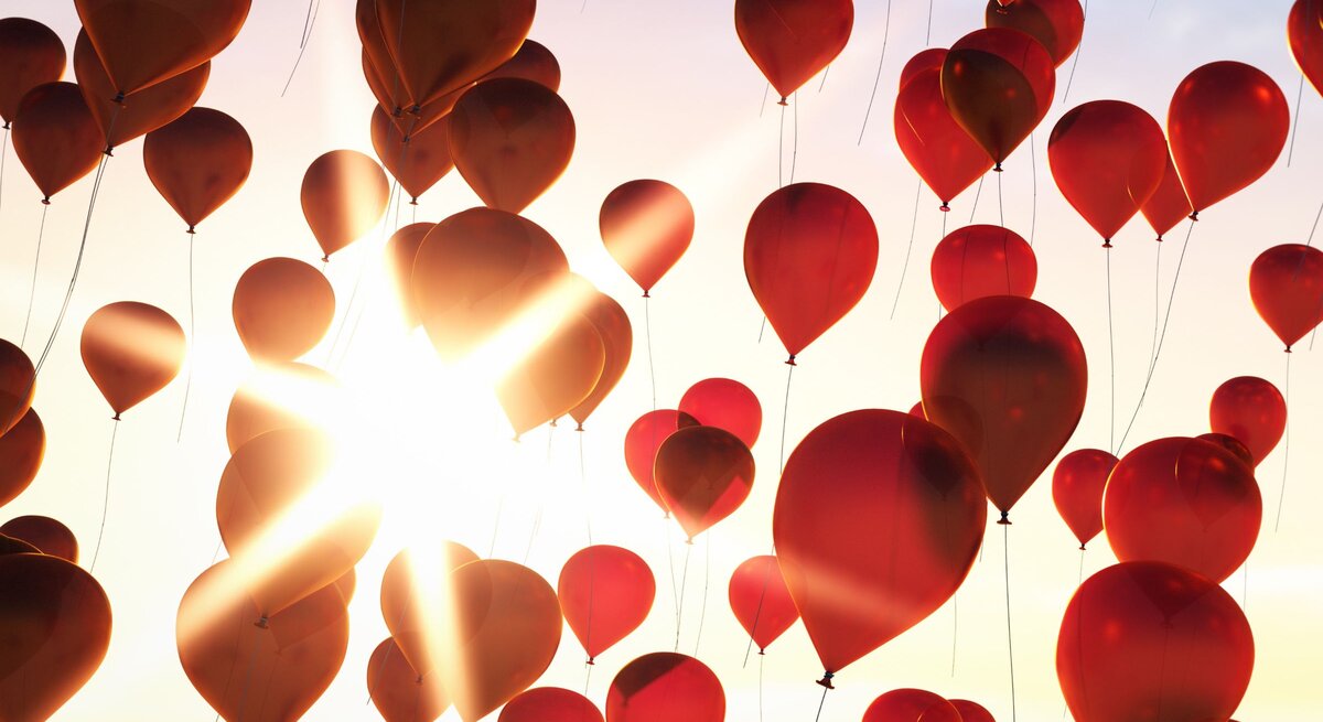 Leuchtend rote Luftballons schweben vor strahlender Sonne in den pastellfarbenen Himmel.