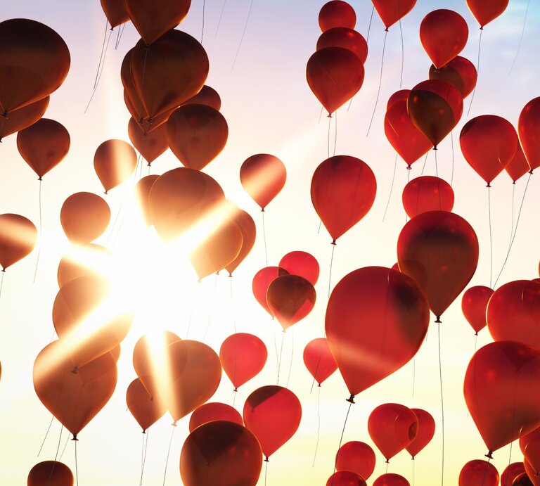Leuchtend rote Luftballons schweben vor strahlender Sonne in den pastellfarbenen Himmel. | © AdobeStock/peterschreiber.media