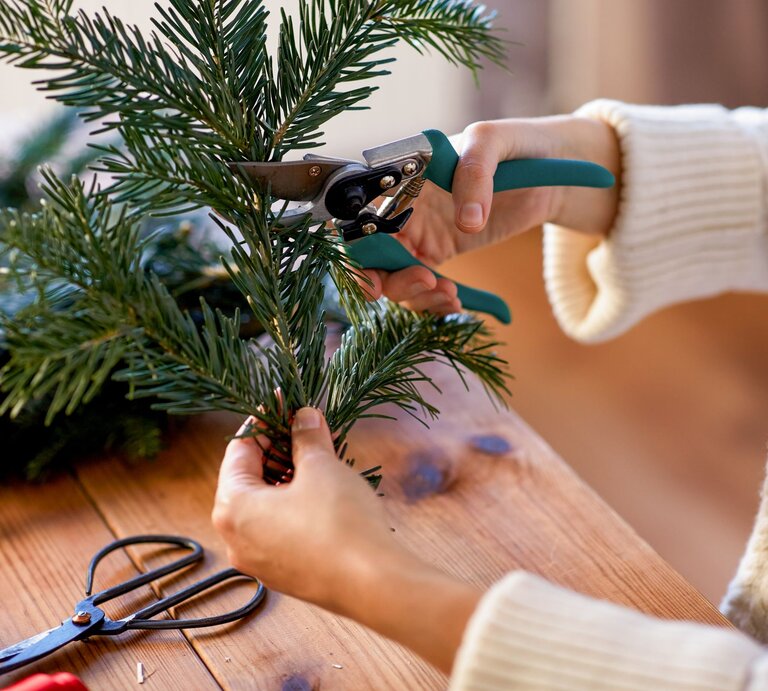 Eine Person in cremefarbenem Strickpullover schneidet mit Gartenschere frische Tannenzweige auf einem Holztisch. | © AdobeStock/Syda Productions