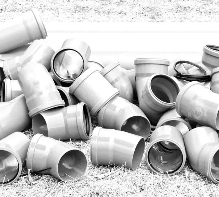Mehrere graue Abflussrohre und HT-Rohre aus Kunststoff liegen ungeordnet auf trockenem, braunem Grasboden. | © AdobeStock/fefufoto