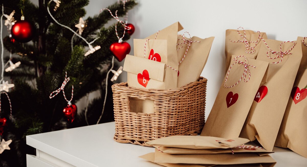 Liebevoll verpackte Adventskalendertüten mit roten Herzen und Zahlen liegen neben einem geschmückten Weihnachtsbaum auf Weiß.
