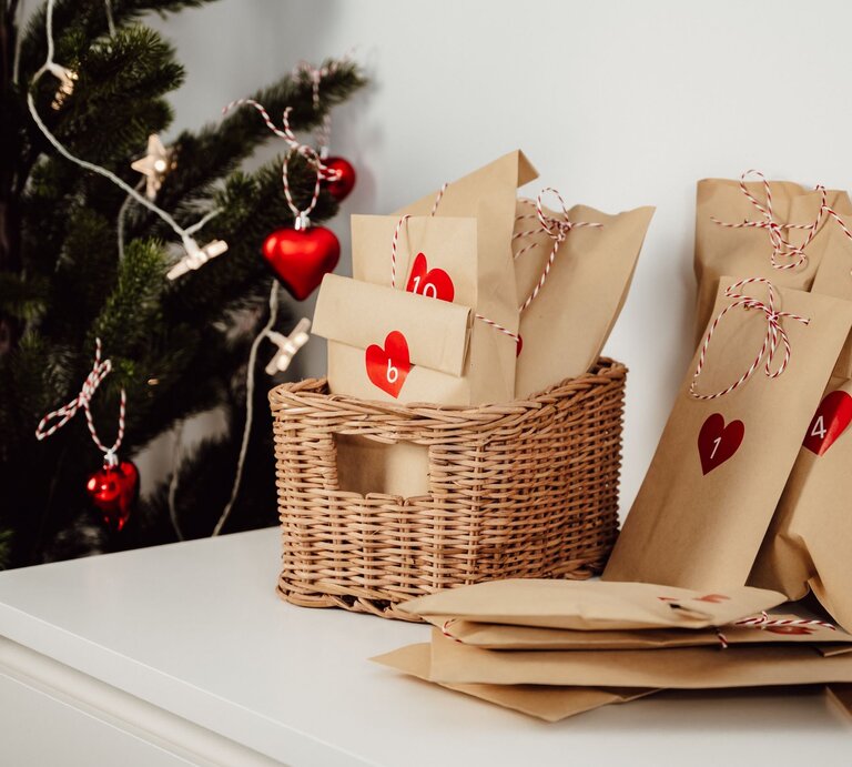 Liebevoll verpackte Adventskalendertüten mit roten Herzen und Zahlen liegen neben einem geschmückten Weihnachtsbaum auf Weiß. | © Shutterstock/piksik