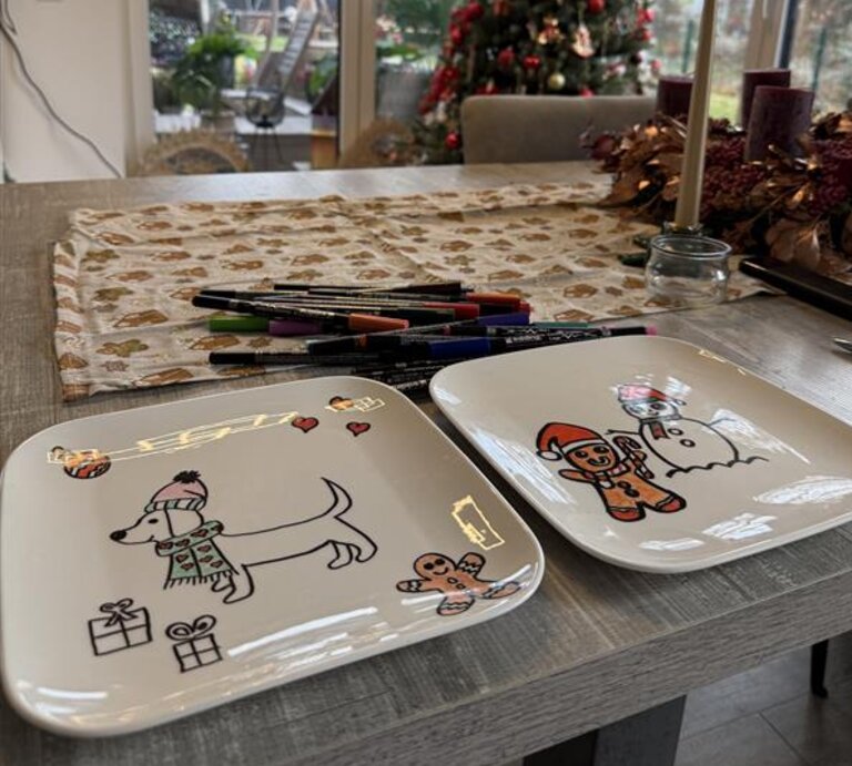Zwei bemalte Weihnachtsteller mit Hund und Lebkuchenmann liegen auf einem Tisch vor festlich geschmücktem Weihnachtsbaum. | © Madeleine Liedke 
