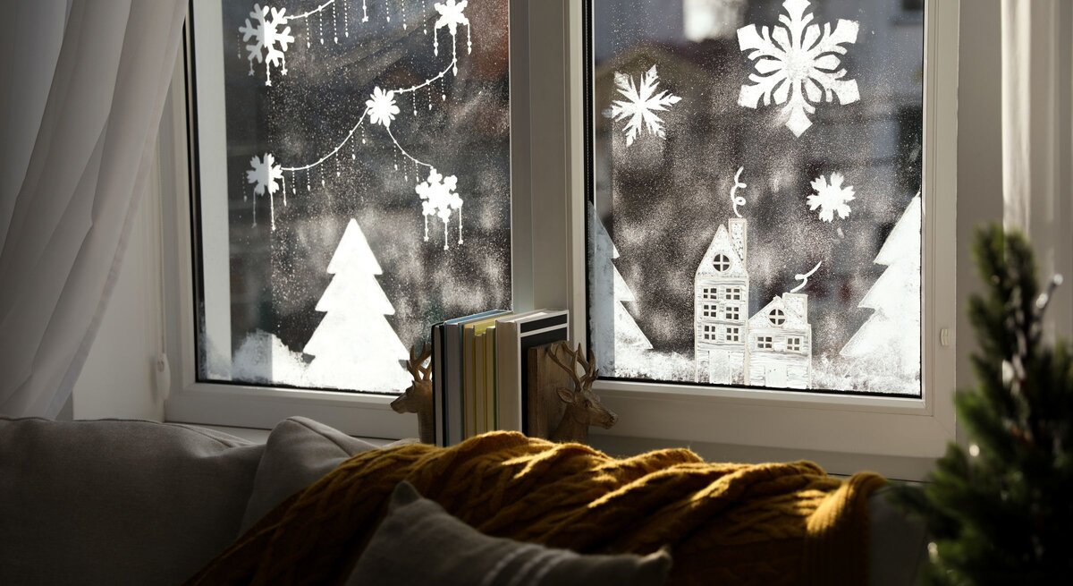 Winterliche Fensterdeko mit Schneeflocken, Tannen und Häuschen, davor Sofa mit Kissen und gelber Strickdecke.