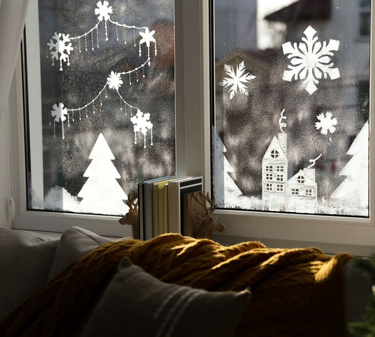 Winterliche Fensterdeko mit Schneeflocken, Tannen und Häuschen, davor Sofa mit Kissen und gelber Strickdecke. | © Shutterstock/New Africa