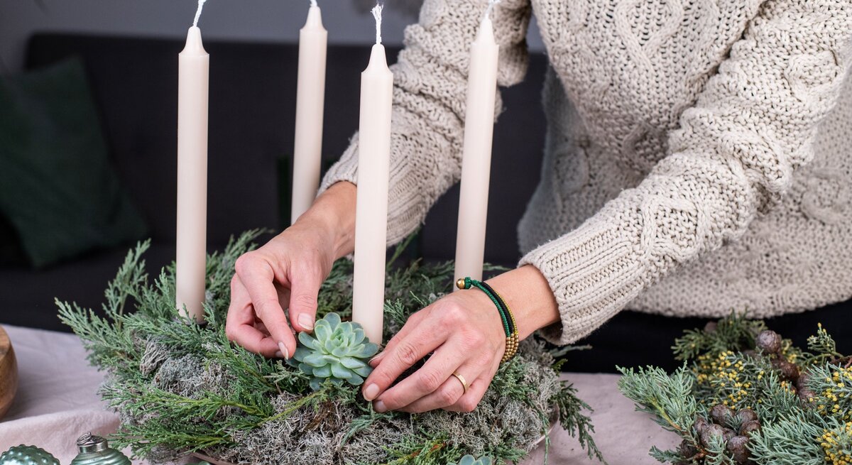 Eine Frau mit beigem Strickpullover dekoriert einen Adventskranz mit Sukkulenten und vier weißen Stabkerzen auf Moosgrund.