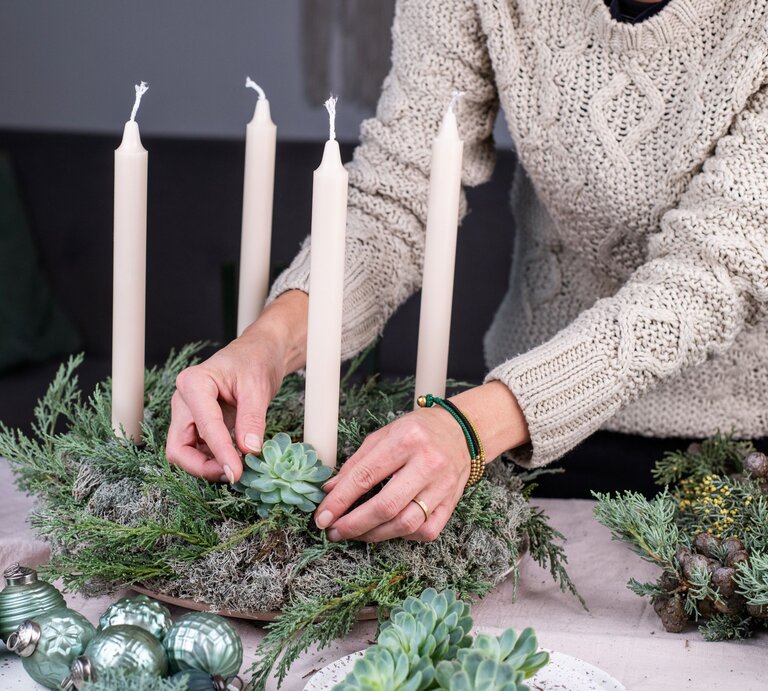 Eine Frau mit beigem Strickpullover dekoriert einen Adventskranz mit Sukkulenten und vier weißen Stabkerzen auf Moosgrund. | © AdobeStock/knipseria