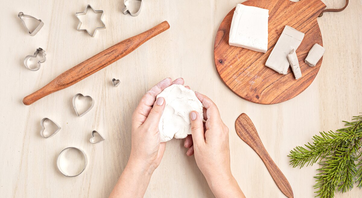 Zwei Hände formen helle Modelliermasse auf hellem Holztisch, umgeben von Ausstechformen, Holzrolle und Tannenast.