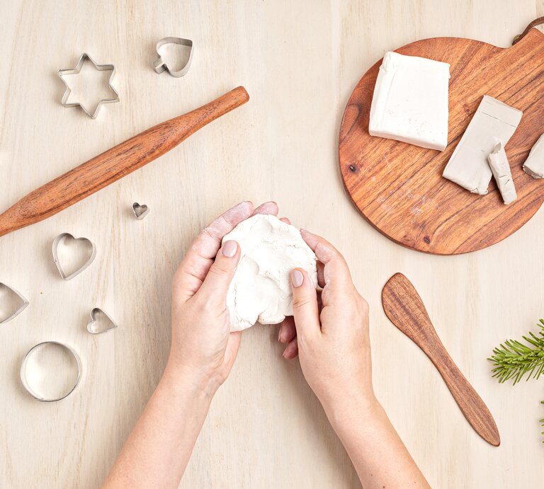 Zwei Hände formen helle Modelliermasse auf hellem Holztisch, umgeben von Ausstechformen, Holzrolle und Tannenast. | © Shutterstock/Netrun78