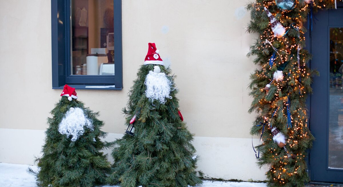 Tannenförmige Weihnachtsfiguren mit Lockenbärten und Zipfelmützen stehen im Schnee neben beleuchtetem Türkranz.
