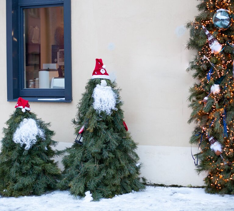 Tannenförmige Weihnachtsfiguren mit Lockenbärten und Zipfelmützen stehen im Schnee neben beleuchtetem Türkranz. | © Shutterstock/Nataliabiruk