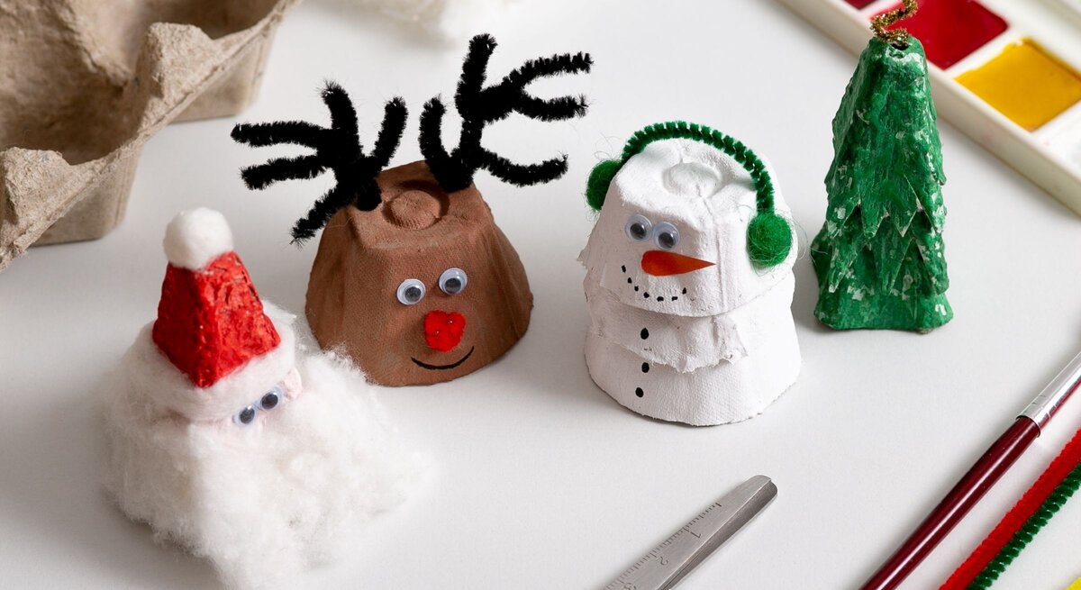 Vier weihnachtliche Figuren aus Eierkarton – Nikolaus, Rentier, Schneemann und Tannenbaum – stehen auf hellem Basteltisch.
